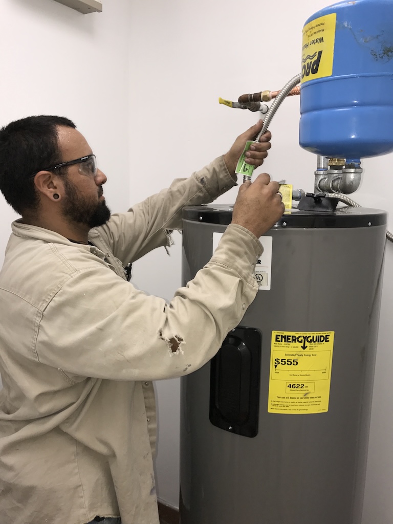 All City Plumbing Technician holds a tube connected to a water heater that he is examining.